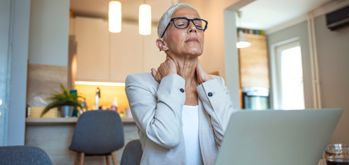 woman with short gray hair and glasses sitting at a desk with a laptop showing signs of discomfort in her neck while working in a modern office environment focusing on workplace wellness for two