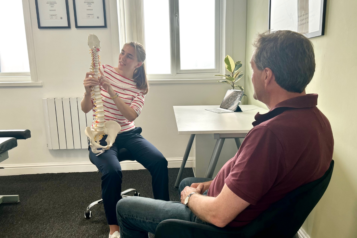 Chiropractor explaining patient's page to them using a spinal anatomical model, taking place in a clinical modern chiropractic clinic
